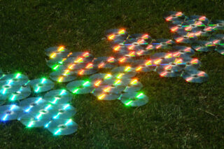 Mehrere CDs sind in überlappenden Gruppen auf dem Gras angeordnet und reflektieren das Sonnenlicht, wodurch helle Flecken in Regenbogenfarben entstehen.