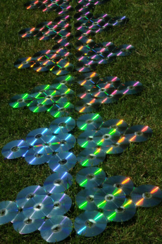 Auf dem Gras angeordnete Reihen von CDs reflektieren im Sonnenlicht regenbogenfarbene Lichtmuster.
