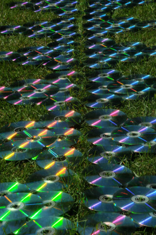 Auf grünem Gras sind Reihen von CDs angeordnet, die regenbogenfarbene Lichtmuster reflektieren.