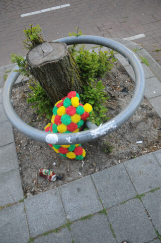 Ein buntes, rundes Plüschtier liegt an einem Baumstumpf auf einem Gehweg, umgeben von einem Metallgeländer. In der Nähe liegt Müll.