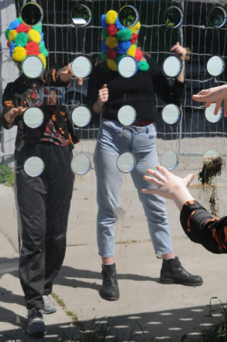 Zwei Personen mit bunten Pompon-Masken stehen hinter einer reflektierenden Oberfläche mit kreisförmigen Ausschnitten, während der Arm einer dritten Person nach ihnen greift.