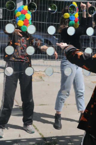 Hinter einem Netz mit runden Spiegeln stehen zwei Personen mit bunten Pompon-Masken; im Vordergrund sind ihre Spiegelbilder und ein Arm zu sehen.