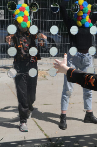 Zwei Personen, deren Gesichter durch bunte Pompons verdeckt sind, stehen hinter einer vertikalen Tafel mit runden Spiegeln, während eine dritte Person nach der Tafel greift.