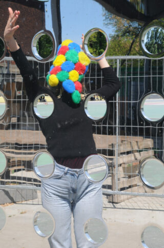 Eine Person mit einer bunten Bommelmütze steht hinter einer reflektierenden Platte mit kreisförmigen Ausschnitten und hebt einen Arm.