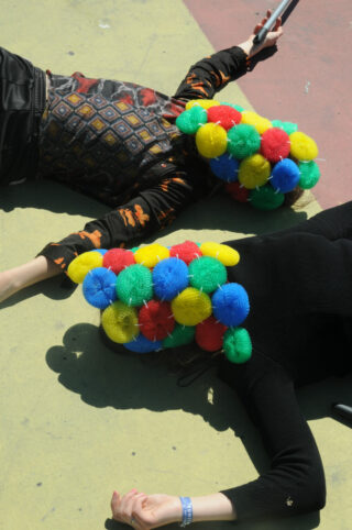 Zwei Personen liegen auf einer Betonoberfläche und tragen jeweils einen Kopfschmuck aus bunten, strukturierten Bällen in Rot, Gelb, Grün und Blau.