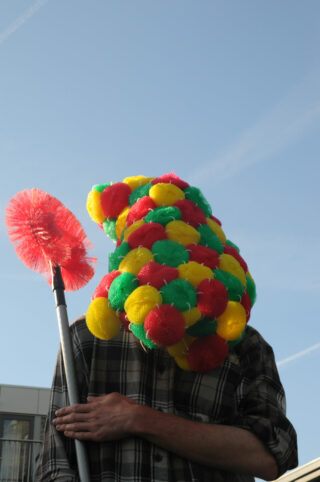 Eine Person mit einer Maske aus bunten Schrubbern hält eine rote Toilettenbürste vor einem klaren Himmel und trägt ein kariertes Hemd.