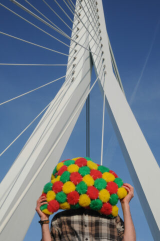 Eine Person hält ein buntes Pompon-Objekt vor ihr Gesicht und steht unter einer modernen weißen Schrägseilbrücke vor einem klaren blauen Himmel.