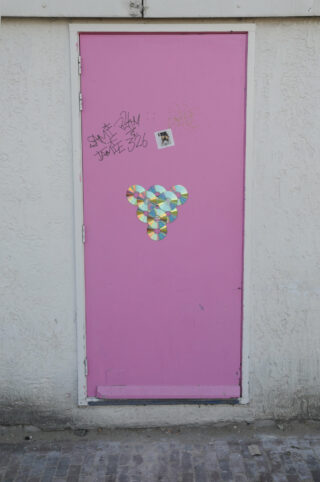 Eine rosa Tür mit Graffiti, einem kleinen Foto und einem Stapel CDs in Dreiecksform ist in eine weiße strukturierte Wand eingelassen.