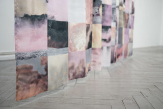 Vor einem hellen Holzboden hängt ein halbtransparenter Stoff mit einem Patchwork aus pastellfarbenen und dunklen aquarellartigen Quadraten.