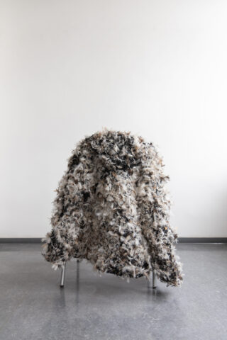Ein strukturierter, zotteliger Mantel in einer Mischung aus Schwarz-, Weiß- und Grautönen hängt über einem Stuhl mit Metallrahmen in einem minimalistischen, leeren Raum mit einer schlichten weißen Wand und einem grauen Boden.