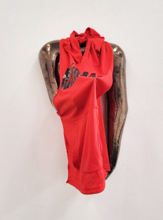 An einer Wand ist eine metallische Skulptur menschlicher Arme angebracht, über die ein rotes Kapuzensweatshirt drapiert ist.