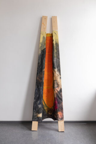 Eine A-förmige Holzkonstruktion stützt einen verwitterten, in Schwarz, Gelb und Orange bemalten Stoff und lehnt an einer schlichten Wand auf einem grauen Boden.
