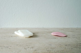 Ein weißes Stück Seife und ein flacher, ovaler rosa Stein liegen auf einer beigen Fläche vor einer schlichten hellen Wand.