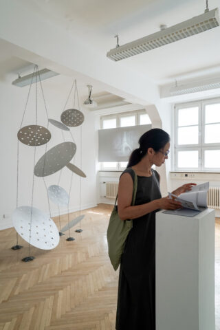 Eine Frau steht und liest ein Buch neben einem weißen Podest in einer Galerie mit hängenden runden Metallskulpturen und großen Fenstern.