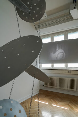 Große perforierte Metallscheiben hängen an Ketten in einem weißen Raum; auf einer Leinwand im Hintergrund wird die Projektion eines klassischen Gemäldes eines menschlichen Gesichts angezeigt.