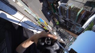 Eine Person lehnt sich mit einer Kamera oder einem anderen Gerät in der Hand über ein Balkongeländer, während weit unten in einem steilen Abwärtswinkel Gebäude und eine Straße zu sehen sind.