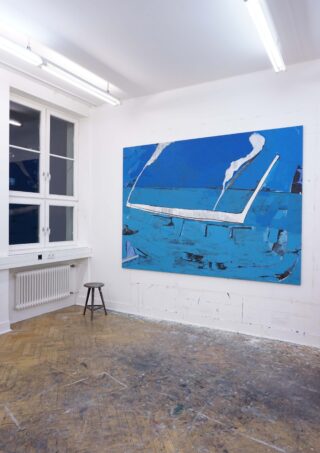 Ein großes blaues abstraktes Gemälde hängt an einer weißen Wand in einem Studio mit Holzboden, weißem Heizkörper, Fenster, Deckenbeleuchtung und einem einzelnen schwarzen Hocker.