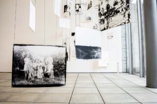 Schwarzweißfotografien hängen an Drähten in einem modernen Galerieraum mit großen Fenstern und gefliestem Boden.