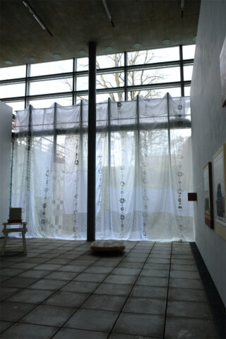 Große, durchsichtige Vorhänge mit Blumenmustern hängen vor hohen Fenstern; eine schwarze Stange, ein rundes Kissen und ein Stuhl stehen im Raum, an dessen weißen Wänden Kunstwerke hängen.