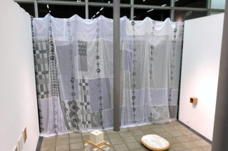 An einer Wand in einem gefliesten Raum hängt ein Patchwork-Vorhang aus verschiedenen weißen und gemusterten Stoffen, auf dem Boden und den Wänden liegen eine Säule und mehrere kleine Gegenstände.