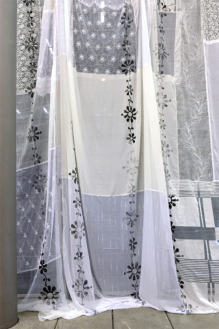 Transparente weiße Vorhänge mit schwarzen Blumen- und geometrischen Mustern hängen vor einer grauen Säule und einer Metallbank.