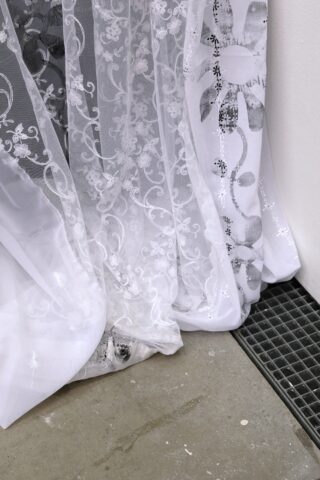 Eine Nahaufnahme von durchsichtigen weißen Vorhängen mit Blumenmustern, die auf einem Betonboden neben einem schwarzen Metallgitter und einer weißen Wand liegen.