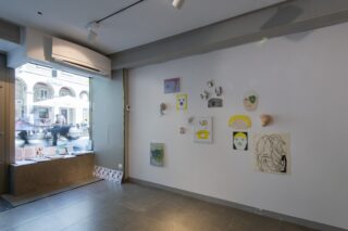 Eine kleine Kunstgalerie mit Zeichnungen, Masken und Kunstwerken aus verschiedenen Medien an einer weißen Wand; durch ein großes Fenster ist eine verschwommene Straßenszene draußen zu sehen.