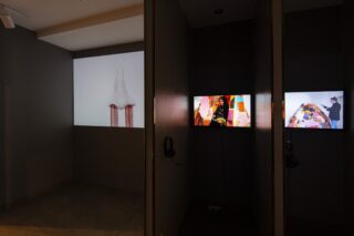 In einem schwach beleuchteten Raum werden auf drei Videobildschirmen verschiedene Kunstbilder gezeigt. An der Wand unter zwei der Bildschirme hängen Kopfhörer.