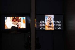 Auf zwei Monitoren wird Kunst angezeigt: Der linke zeigt einen Mann, der zwischen bunten Objekten sitzt, der rechte zeigt den wiederholten Text „Ich habe keine Freunde“ vor einem unscharfen Hintergrund.