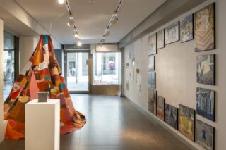 Innenraum einer Kunstgalerie mit einem farbenfrohen Patchwork-Tipi auf der linken Seite und gerahmten Kunstwerken, die in einem Raster an der rechten Wand angeordnet sind, mit großen Fenstern, die natürliches Licht hereinlassen.
