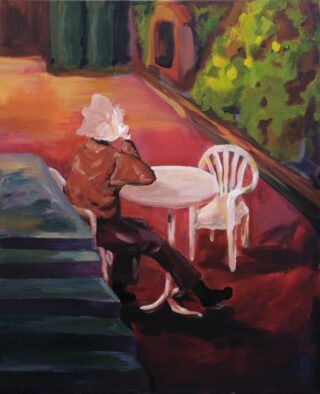 Eine Person mit weißem Haar sitzt im Freien an einem runden weißen Tisch und telefoniert. Im Hintergrund sind zwei leere weiße Stühle und Grünpflanzen zu sehen.