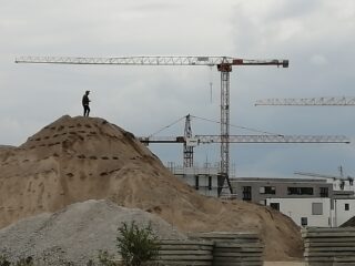 Eine Person steht auf einem Sandhügel auf einer Baustelle, im Hintergrund sind unter einem bewölkten Himmel Kräne und teilweise errichtete Gebäude zu sehen.