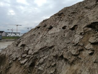 Großer Erdhaufen auf einer Baustelle mit Kränen und teilweise fertiggestellten Gebäuden im Hintergrund unter einem bewölkten Himmel.