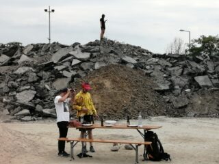 Drei Personen im Freien: zwei stehen mit Getränken und Essen an einem Tisch vor Trümmern und eine steht allein auf einem Felshügel im Hintergrund.