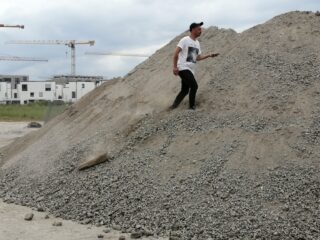 Ein Mann in einem weißen T-Shirt und einer schwarzen Hose geht auf einer Baustelle einen großen Kieshaufen entlang, im Hintergrund sind Kräne und Gebäude zu sehen.