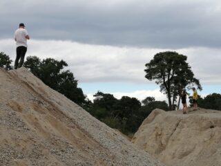 Ein Mann steht auf einem Kieshügel und macht ein Foto von drei Personen, darunter ein Kind, die auf einem anderen Hügel stehen, mit Bäumen und Wolken im Hintergrund.