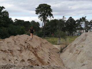 Eine Person in schwarzem Outfit läuft im Freien auf einem Erdhügel, im Hintergrund sind Bäume und ein bewölkter Himmel zu sehen.