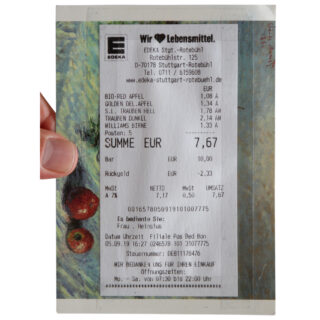 Eine Hand hält einen deutschen Supermarktbeleg für Obst im Gesamtwert von 7,67 Euro, der auf einer bunten Postkarte mit Obstmotiven liegt.