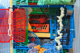 Eine farbenfrohe Anordnung von Plastikkisten, Netzpaneelen und Schaumstoffstrukturen mit verschiedenen Formen und Farben, die vor einem blauen Gitterhintergrund übereinandergelegt sind.