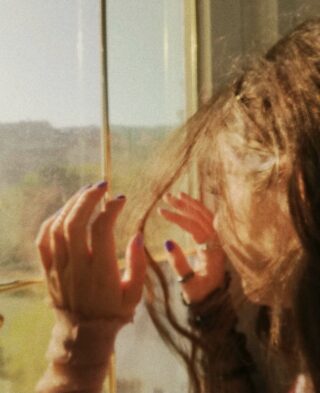 Eine Person mit langen Haaren und lackierten Nägeln hält ihre Hände vor ein sonnenbeschienenes Fenster und blickt nach draußen. Das Bild ist sanft fokussiert und wird von natürlichem Licht erhellt.