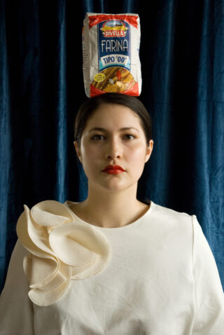 Eine Frau in einer weißen Bluse mit Rüschendetail steht vor einem dunkelblauen Hintergrund und balanciert einen Sack Divella Farina Tipo '00'-Mehl auf ihrem Kopf.