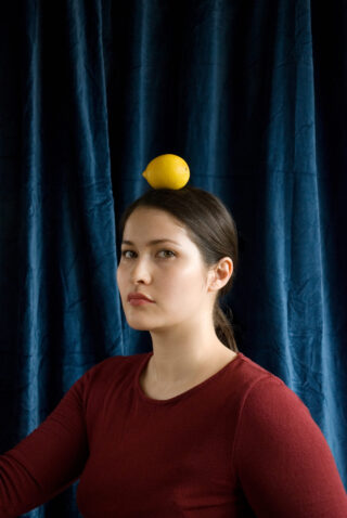 Eine Frau mit dunklem Haar in einem roten Hemd sitzt vor einem blauen Vorhang und balanciert eine Zitrone auf ihrem Kopf.