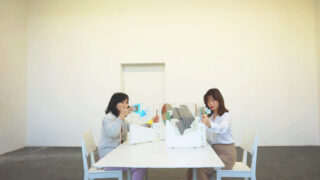 Zwei Frauen sitzen sich an einem weißen Tisch mit Trennwänden gegenüber und halten jeweils ein Glas Wasser in einem minimalistischen Raum mit einer schlichten cremefarbenen Wand.