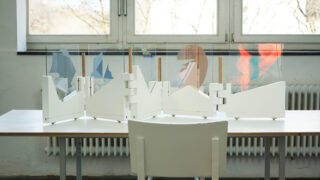 Ein weißer Stuhl und ein Tisch mit einer Trennwand aus farbenfrohen abstrakten Formen stehen vor Fenstern, von denen aus man Bäume sehen kann.