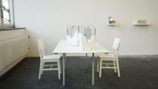 Ein weißer Tisch mit zwei weißen Stühlen steht in einem minimalistischen Raum. Der Tisch ist mit abstrakten Glastrennwänden versehen, und an der Rückwand ist ein Regal mit Büchern und Kunstwerken angebracht.