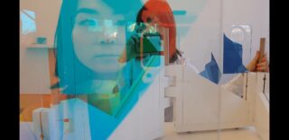 In einer Glasscheibe mit blauen und türkisfarbenen geometrischen Formen spiegelt sich eine Person, die ein Telefon in einem modernen, minimalistischen Raum mit weißen Strukturen hält.