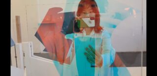 Eine Person in einer weißen Jacke macht ein Selfie vor einer Glaskunstinstallation mit überlappenden blauen und roten Formen.