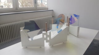 Eine geometrische Skulptur mit farbigen Glasplatten und weißen Sockeln steht auf einem weißen Tisch neben einem Fenster, unter dem sich ein Heizkörper befindet.