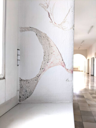 Eine Eckwand in der Nähe eines Fensters weist eine zarte, abstrakte Zeichnung mit Punktmustern auf, in einem hellen Flur mit gefliesten Böden und gewölbten Türen.