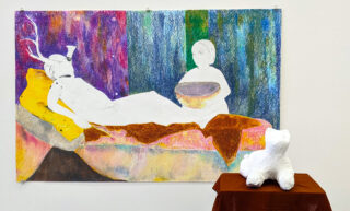 Ein farbenfrohes Kunstwerk zeigt eine liegende, teilweise umrissene Figur mit Geweih und eine andere, die eine Schüssel hält; davor sitzt auf einem kleinen, braun bedeckten Tisch ein weißes, geformtes Tier.